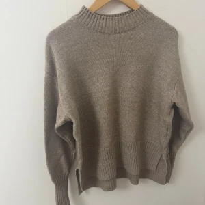 Beige stickad tröja med ribbade detaljer - En beige stickad tröja med hög ribbad krage och breda ribbade muddar vid ärmslut och nederkant. Tröjan har en avslappnad passform och slitsar i sidorna, perfekt för lager-på-lager under kyliga dagar.