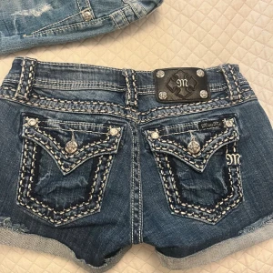 Miss Me jeansshorts med broderi - Snygga blå jeansshorts från Miss Me med kraftiga kontrastsömmar, dekorativa bakfickor med broderi och blingiga knappar. Klassisk femficksmodell med låg midja och uppvikta benslut. Perfekta för dig som gillar detaljer och statement-plagg.