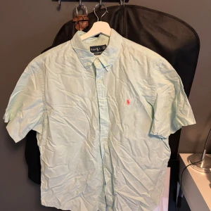 Ljusblå kortärmad skjorta Ralph Lauren - Fräsch ljusblå skjorta från Ralph Lauren i klassisk fit, kortärmad med button-down krage och rosa broderad logga på bröstet. Skjortan är i bomull och har knappar framtill. Perfekt för en chill och stilren look.