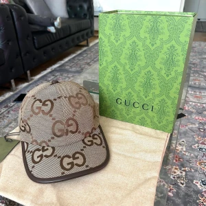 Gucci keps med GG-mönster i beige och brunt - Snygg keps från Gucci i beige och brunt med det klassiska GG-mönstret över hela kepsen. Kepsen har justerbar rem baktill i mörkbrunt skinn och bruna detaljer längs skärmen. Tillverkad i canvasmaterial med exklusiv känsla, perfekt för dig som gillar lyxiga accessoarer.
