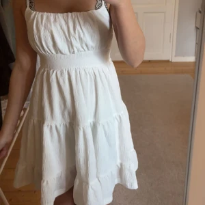 Vit somrig kortklänning med volanger - Superfin vit klänning med smala axelband och markerad midja. Klänningen har en luftig, volangig kjol och lätt rynkade detaljer upptill. Perfekt för varma dagar och har en fräsch, enkel look som känns både trendig och tidlös. Aldrig använd