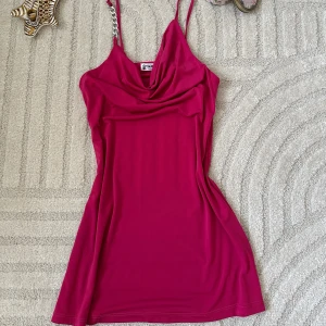 Rosa kortklänning med kedjedetalj - Snygg rosa kortklänning från Soie med tunna axelband, varav ett har en cool kedjedetalj. Klänningen har draperad urringning och är figurnära med slinky känsla. Perfekt för dig som vill sticka ut med en färgstark look.