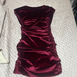 Vinröd sammetsklänning med rynk och öppen mage  - Supersnygg vinröd kort sammetsklänning med rynkade sidor och axelvaddar. Klänningen har en tight passform och är ärmlös, perfekt för dig som vill sticka ut. Materialet är glansigt och mjukt, vilket ger en lyxig känsla.