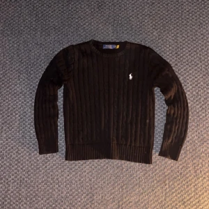 Svart stickad tröja från Polo Ralph Lauren - Kabelstickad svart tröja från Polo Ralph Lauren med klassisk rund halsringning och vit broderad logga på bröstet. Tröjan har långa ärmar och ribbade muddar för en snygg passform. Perfekt för lager-på-lager under kyliga dagar.