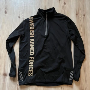 Svart half zip tröja South West M - Svart långärmad tröja från South West med halv dragkedja och tryck 'Swedish Armed Forces' i beige på både fram- och baksida. Tröjan har hög krage och reflekterande detaljer. Perfekt för träning eller chill dagar.