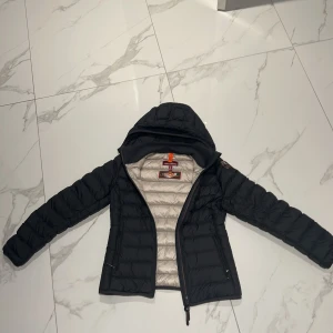 Svart dunjacka Parajumpers S - Svart dunjacka från Parajumpers i storlek S. Jackan är superlätt, har quiltad design, huva och dragkedja framtill. Insidan är ljusbeige och jackan har klassisk PJS-patch på ärmen. Perfekt för kyliga dagar när du vill ha både stil och komfort. Jackan har inga hål eller slitningar.