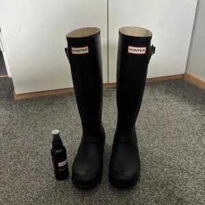 Svarta höga stövlar från Hunter - Säljer ett par klassiska svarta höga stövlar från Hunter med justerbart spänne på sidan och rund tå. Tillverkade i slitstarkt syntetmaterial och är riktigt snygga och trendiga boots. Insidan är ljus med diskret mönster. En flaska Hunter Boot Shine medföljer, hör av er vid frågor😊