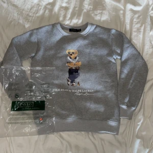 Grå Polo Bear sweatshirt från Ralph Lauren - Grå sweatshirt från Ralph Lauren med ikoniska Polo Bear-trycket framtill. Tröjan har rund hals, långa ärmar och ribbade muddar. Perfekt för dig som gillar klassisk streetstyle med en twist. Mjuk insida och snygg passform.