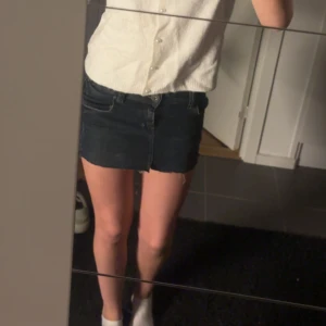 Mörk jeanskjol med rå kant - Säljer en mörkblå jeanskjol med rå, fransig kant nertill. Kjolen har klassisk femficksdesign, knapp och dragkedja framtill samt bakfickor med dekorativa sömmar. Perfekt för dig som gillar en enkel och stilren look.