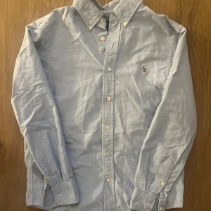 Ljusblå skjorta Ralph Lauren - Klassisk ljusblå skjorta från Ralph Lauren med broderad logga på bröstet. Skjortan har button-down krage, långa ärmar och stängs med vita knappar framtill. Tillverkad i bomull och passar perfekt till en clean och stilren look.                    Öppen för frågor och bud!