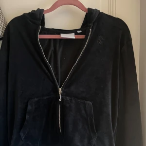 Svart velourhoodie från Juicy Couture - Svart hoodie i mjuk velour från Juicy Couture med dragkedja och huva. Klassisk broderad JC-logga på bröstet och ficka framtill. Dragkedjan har en snygg J-detalj. Perfekt för en chill och trendig look.