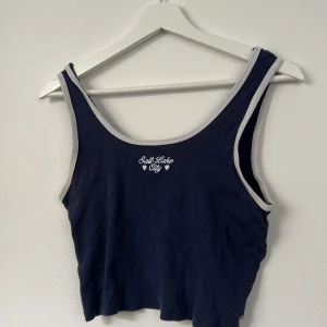 Mörkblå croppad linnetopp Pull&Bear - Croppad mörkblå linnetopp från Pull&Bear med ribbad struktur och ljusgrå kantband. Tryck med texten 'Salt Lake City' och små hjärtan framtill. Perfekt för en chill och sportig look.