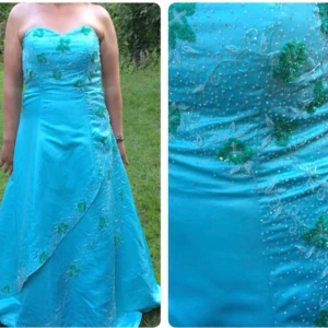 Turkos axelbandslös balklänning - Turkos långklänning med axelbandslös, hjärtformad överdel. Klänningen har gröna blommiga broderier och små pärlor över hela livet och kjolen. Materialet är glansigt och ger en lyxig känsla, perfekt för bal eller speciella tillfällen.