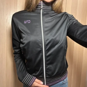 Svart track jacket från CRW med lila detaljer - Svart track jacket/kofta från CRW med lila och vita ränder på krage och muddar. #TrackJacket #Retro #Sportkläder #Jacka #Mode #Stil #Vintage #CRW #Avslappnad #Kläder #Fashion #Outfit #LilaDetaljer #SilverDragkedja #ridingwear