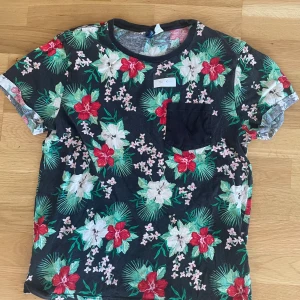 Blommig svart t-shirt med ficka - Svart t-shirt från Cubus med färgglatt blommigt mönster i rött, vitt och grönt. Har en svart bröstficka och rund hals. Perfekt för dig som gillar tropiska vibes och vill sticka ut med en cool print. Materialet känns mjukt och skönt mot huden.