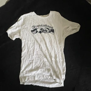 Vit t-shirt från Arontattoos - Vit t-shirt från Arontattoos med svart tryck framtill och liten logga i nacken. Klassisk rund hals och korta ärmar. Skön och enkel modell i bomull, perfekt till jeans eller shorts.