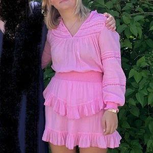 Rosa volangklänning från Neo Noir - Supersöt rosa kortklänning från Neo Noir med långa ärmar, v-ringning och smock i midjan. Klänningen har flera volangdetaljer och är gjord i ett tunt, luftigt bomullstyg. 