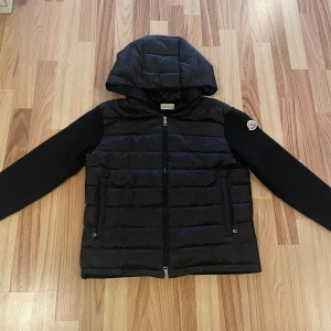 Svart Moncler pufferjacka med huva - Svart pufferjacka från Moncler med quiltad front, stickade ärmar och huva. Den är väldigt skön o helt oanvänd då den inte passade mig, skulle säga att den passar 170-180cm. Pris kan diskuteras