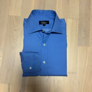 Blå skjorta från Oscar Jacobson - Klassisk blå skjorta från Oscar Jacobson tillverkad i twofold bomull med fina detaljer och en lyxig känsla. Skjortan har lång ärm, eleganta knappar och en diskret broderad logga vid nederkanten.  Hör gärna av dig vid frågor, mått eller om du vill ha fler bilder.