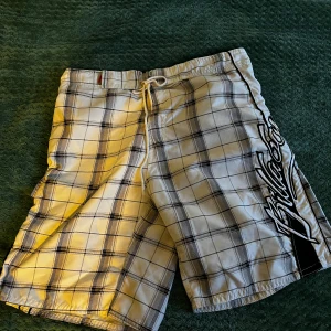 Rutiga badshorts från Billabong - Snygga vita och svarta rutiga badshorts från Billabong med snörning i midjan och stort tryck längs benet. Perfekta för stranden eller poolen, klassisk surfstil och bekväm passform. Kapsylöppnare i fickan