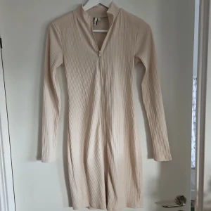 Beige ribbad kortklänning med zip - Snygg beige kortklänning med ribbad struktur och lång ärm. Klänningen har en dragkedja framtill och står ut med sin stilrena look. Perfekt för dig som gillar minimalistisk design och mjuka material.