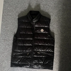 Svart dunväst från Moncler - Svart dunväst från Moncler med klassisk quiltad design och hög krage. Västen har dragkedja framtill, två fickor med dragkedja och Moncler-logga på bröstet. Fylld med vitt gåsdun för extra värme och lätt känsla. Perfekt för lager-på-lager under kyliga dagar.