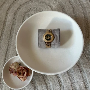 Guldig klocka från Michael Kors - Snygg klocka från Michael Kors med guldig länk och rund urtavla. Urtavlan är svart med stora MK-bokstäver och glittriga detaljer vid siffrorna. Klockan har en stilren och lyxig känsla, perfekt för dig som gillar statement-accessoarer.