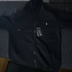 Svart hoodie från Polo Ralph Lauren - Svart hoodie från Polo Ralph Lauren med dragkedja framtill och klassisk vit logga på bröstet. Tröjan har huva med snörning och känguruficka. Perfekt för en chill och stilren look. Pris kan diskuteras!