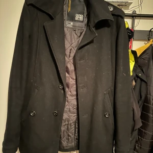 Svart rock från Jack & Jones, stl S - Säljer en svart rock från Jack & Jones i storlek S. Jackan har klassisk krage, knäppning med knappar och två fickor framtill. Insidan är quiltad för extra värme och den har en stilren look som funkar till många outfits. Kan ev behövas en tvätt då ser ut som de finns något bak på jackan.