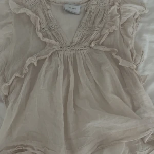 Beige volangblus från Neo Noir - Supersöt beige blus från Neo Noir med volangdetaljer och v-ringning. Tunn och luftig med smockade partier upptill och längs axlarna. Perfekt för dig som gillar romantisk och bohemisk stil.