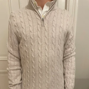 John Henric kabelstickad quarterzip - Kabelstickad beige tröja med hög krage och half zip-dragkedja. Tröjan har lång ärm och klassisk kabelmönster, perfekt för lager-på-lager. Passar till det allra mesta, bland annat jeans och linnebyxor. 