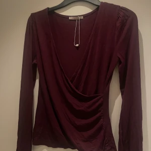 Vinröd omlottopp från Anna Field - Snygg vinröd omlottopp från Anna Field med lång ärm och markerad axel. Toppens omlottdesign ger en schysst v-ringning och smickrande passform. Tillverkad i mjuk bomullsmix, perfekt för dig som gillar stilrena och bekväma plagg.