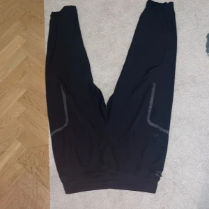 Svarta träningsbyxor med reflexdetaljer - Svarta träningsbyxor med elastisk midja under armour och snygga reflexdetaljer längs benen. Perfekta för löpning eller gymmet, med en loose passform som ger skön rörelsefrihet. Tillverkade i ett lätt syntetmaterial som andas.