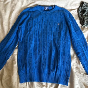 Blå kabelstickad tröja från Gant - Kabelstickad blå tröja från Gant med rund halsringning och diskret logga broderad på bröstet. Tröjan har långa ärmar och ribbade muddar vid ärmslut och nederkant. Perfekt för dig som gillar klassisk och stilren look. Ny pris 1900