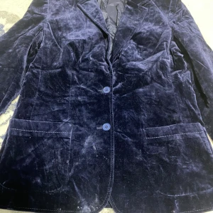 Mörkblå sammetskavaj i storlek 40 - Snygg mörkblå kavaj i mjuk sammet med två knappar framtill och klassisk krage. Fodrad insida och två stora fickor framtill. Perfekt för dig som gillar en elegant och cool stil med lite extra lyxkänsla.