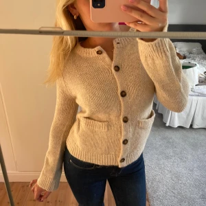 Beige stickad kofta med knappar - Mysig beige stickad kofta med rund hals och stora bruna knappar framtill. Koftan har två små fickor och ribbade muddar vid ärmslut och nederkant. Perfekt för lager-på-lager och chill dagar.