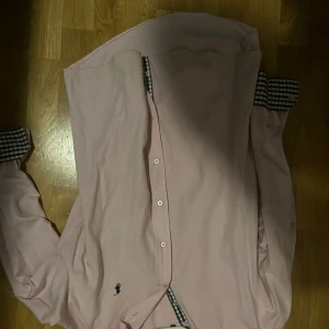 Rosa skjorta från Polo Ralph Lauren - Rosa skjorta från Polo Ralph Lauren med klassisk krage, knappar framtill och långärmade ärmar. Snygga rutiga detaljer i svart och vitt på insidan av krage och manschetter. Broderad logga på bröstet. Skön bomullskvalitet.