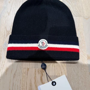 Svart Moncler mössa  - Svart ribbstickad mössa från Moncler med klassiska röda, vita och blå ränder på uppviket. Mössan har Moncler-logga framtill och är tillverkad i mjukt ullmaterial. Perfekt accessoar för att hålla stilen kallare dagar.