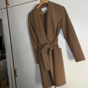 Brun kappa från Vila med bälte - Stilren brun kappa från Vila i klassisk modell med knytbälte i midjan och långa ärmar. Jackan har en öppen front och är tillverkad i ett mjukt material som ger en snygg och clean look. Perfekt för dig som gillar minimalistisk stil.