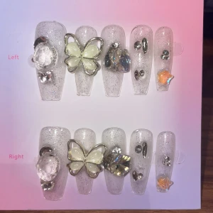 S - Glittriga lösnaglar med fjäril och stenar - Säljer ett set med långa, transparenta lösnaglar med silverglitter, dekorerade med stora kristaller, 3D-fjäril i pärlemor och små stenar. Naglarna har en cool mix av bling och söt design, perfekta för att sticka ut.