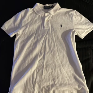 Vit pikétröja Polo Ralph Lauren slim fit - Vit pikétröja från Polo Ralph Lauren i slim fit-modell. Klassisk krage, korta ärmar och knäppning framtill. Ikoniska broderade loggan i marinblått på bröstet. Tillverkad i mjuk bomull, perfekt för en clean och stilren look.