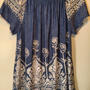 Blåvit kortklänning med blommigt mönster - Supersöt blå kortklänning med vitt blommigt och ornamenterat mönster. Klänningen har korta ärmar och en lös, flowy passform. Materialet känns som bomull och har en denimliknande look. Perfekt för dig som gillar boho-vibes och unika detaljer.