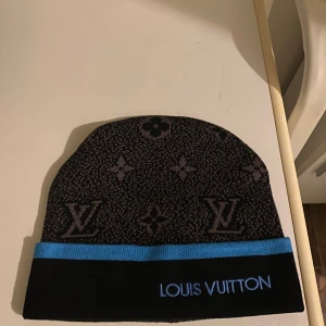 Louis Vuitton svart mössa med blå kant - Snygg svart mössa från Louis Vuitton med klassiskt monogrammönster i grått och en blå kant längst ner. Märkets logga är broderad framtill i blått. Mössan är stickad och har en mjuk, rundad form som sitter skönt på huvudet. Perfekt för dig som gillar exklusiva accessoarer.