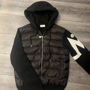 Moncler Cardigan - Säljer en svart cardigan jacka från Moncler med vadderad framsida och huva. Jackan har dragkedja två fickor fram och en vit detalj på ena armen. Jackan har NFC på loggan och tag som man kan scanna. Inte använd. Passar som S-M