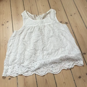 Vit ärmlös linnetopp med spetskant - Söt vit ärmlös linnetopp med broderad spetskant nertill och rund halsringning. Toppen har en synlig dragkedja i ryggen som ger en cool detalj. Perfekt för varma dagar och enkel att styla till jeans eller kjol.