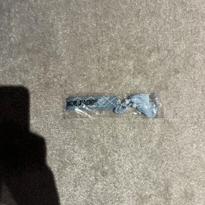 Blått armband med svart text - Snyggt blått armband med texten 'J'Adior' och 'Christian' broderat i svart. Armbandet har ett diskret rutmönster och avslutas med en flätad knytning och tofsar. Perfekt accessoar för att lyfta din outfit med lyxig vibe.