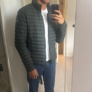 Mörkgrön dunjacka från Colmar - Snygg mörkgrön dunjacka från Colmar med quiltad design och dragkedja framtill. Jackan har hög krage, lång ärm och en clean look med diskret logga på ärmen. Perfekt för dig som vill ha en lätt och stilren jacka till höst och vinter.