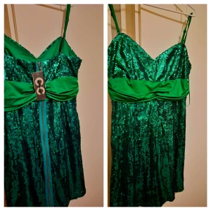 Grön paljettklänning med smala axelband - Glittrig grön kortklänning från GC med smala axelband och markerad midja i satin. Klänningen har v-ringad urringning, dragkedja bak och är täckt av paljetter som ger en riktigt festlig look. Perfekt för dig som vill sticka ut och glänsa.