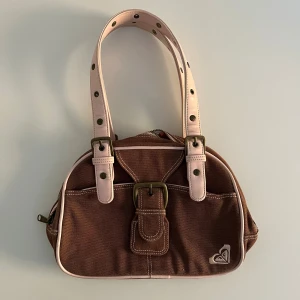 Brun handväska från Quicksilver med rosa detaljer - Snygg brun handväska från Quicksilver/Roxy med ljusrosa handtag och detaljer. Väskan har en  spänndetalj framtill, dragkedja upptill och ett litet broderat Roxy-logo i hörnet. Tillverkad i bomullstyg.