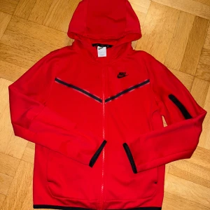  Nike tech ziphoodie junior XL, röd - Nike tech  ziphoodie röd  i storlek junior XL(158-170), med svarta detaljer och dragkedja framtill. Jackan har huva, svarta muddar och en sportig look med svarta ränder över bröstet. Perfekt för dig som gillar streetwear och sportig stil.